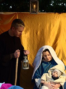 In einer lebendigen Krippe sieht man Josef neben Maria, mit ihren Sohn Jesus im Arm, stehen. Davor scharen sich Kinder die dem Krippenspiel beiwohnen.