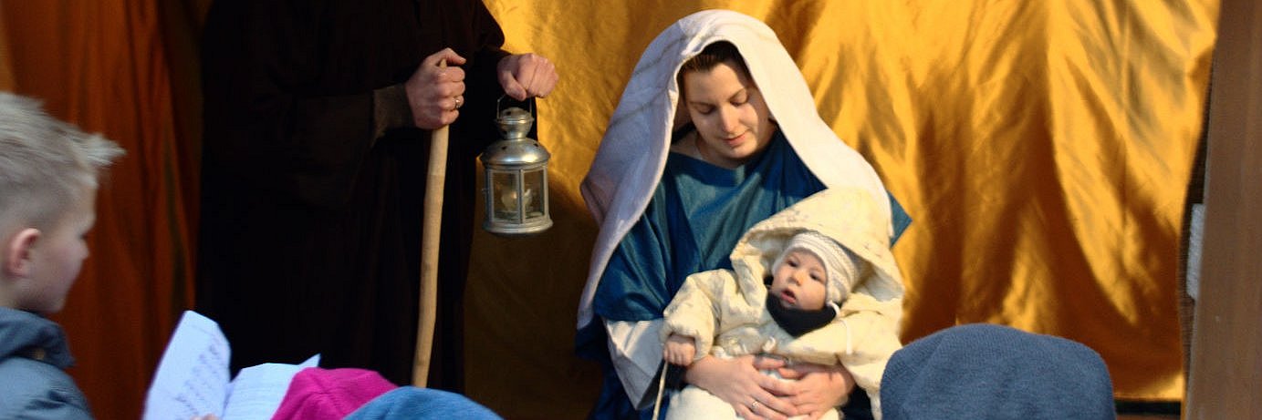 In einer lebendigen Krippe sieht man Josef neben Maria, mit ihren Sohn Jesus im Arm, stehen. Davor scharen sich Kinder die dem Krippenspiel beiwohnen.