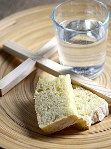 Holzteller auf dem ein einfaches Holzkreuz, ein Stück trockenes Brot und ein Glas Wasser liegen