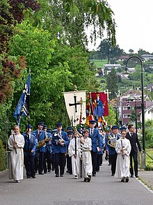Ein Prozessionszug mit Messdienern, Fahnen, Spielmannszug und Gemeinde zieht an Fronleichnam durch die Straßen eines Ortes