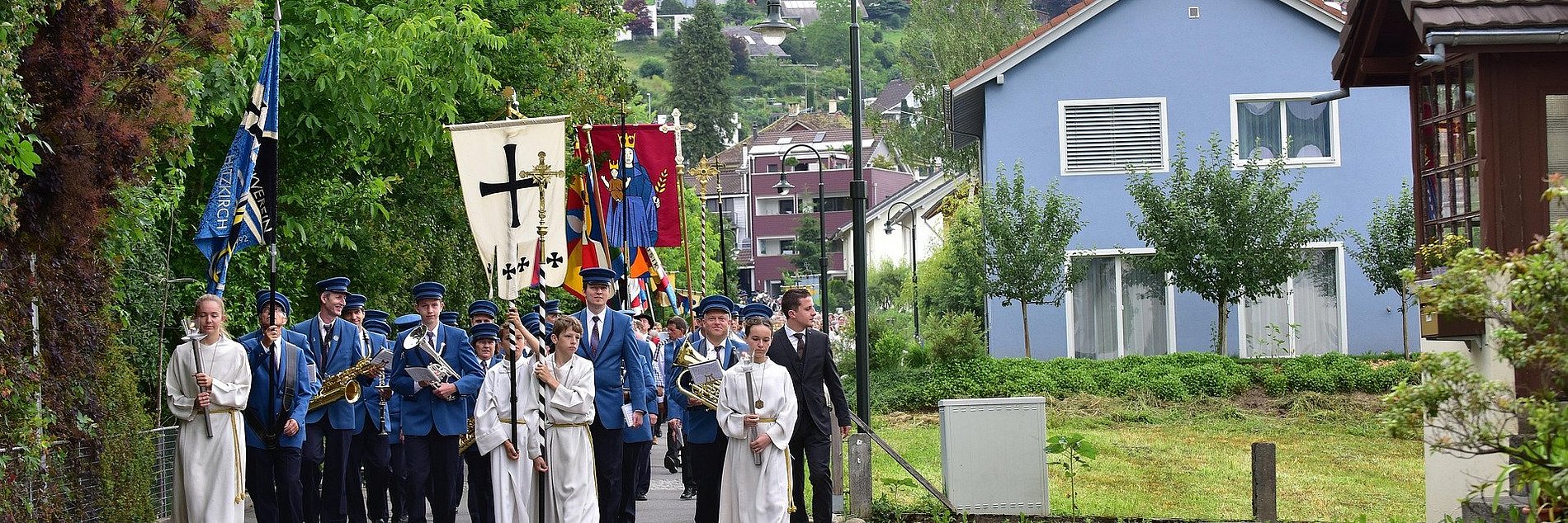 Ein Prozessionszug mit Messdienern, Fahnen, Spielmannszug und Gemeinde zieht an Fronleichnam durch die Straßen eines Ortes